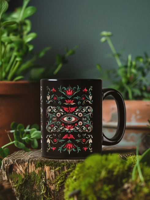 A black ceramic mug featuring a symmetrical folk art design with a central mystic eye motif in turquoise and coral. The design includes ornate scrollwork, red tulip-like flowers, sage green leaves, and cream flourishes arranged in a decorative pattern. The mug has a glossy finish with a C-shaped handle.
